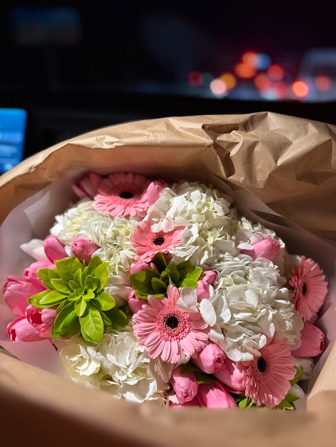 A bouquet of fresh flowers consisting of white hydrangeas, pink gerbera daisies, and small pink buds, arranged with vibrant green foliage. The flowers are wrapped in light brown kraft paper and placed