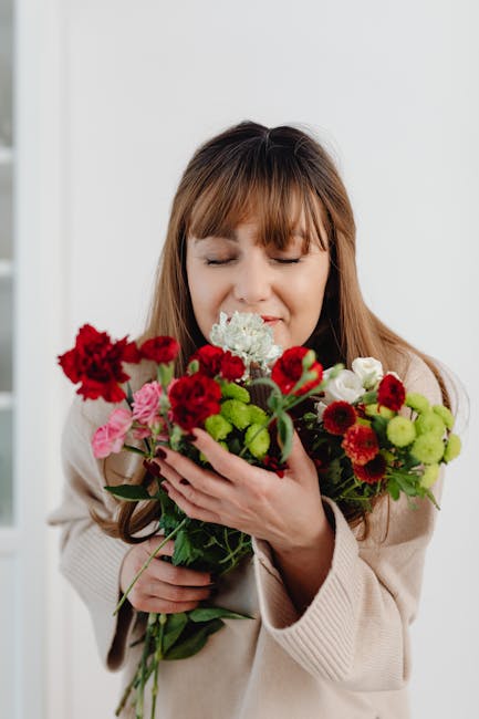 A woman with light brown hair and bangs, dressed in a beige sweater, gently holds a vibrant bouquet of fresh flowers close to her face with her eyes closed and a soft smile. The bouquet includes red c