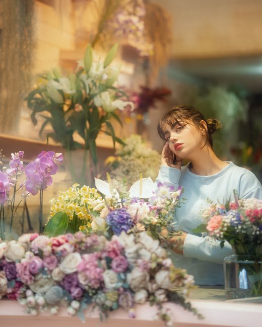 A young woman with shoulder-length brown hair and bangs, wearing a light blue sweater, is seen in a floral shop surrounded by colorful flower arrangements and bouquets. She appears to be contemplating