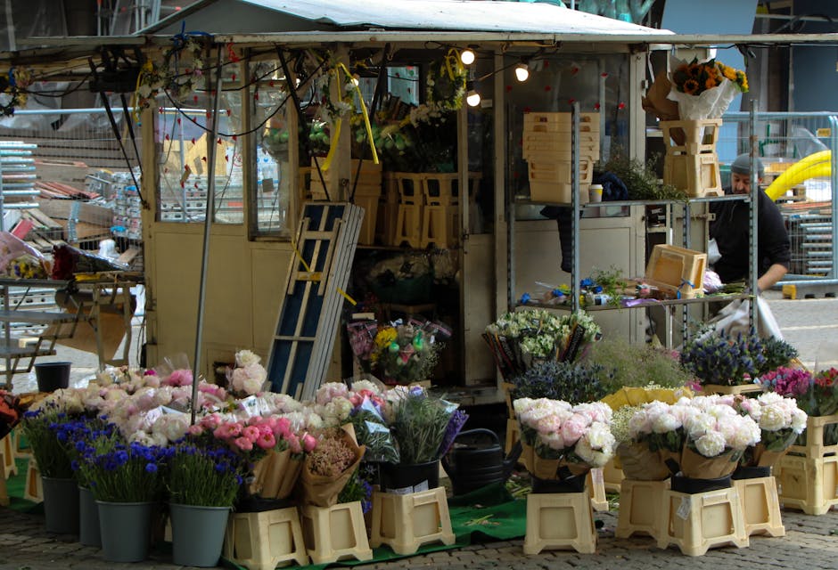 A vibrant outdoor flower stall showcasing an array of fresh floral arrangements and bouquets. The display includes pink, white, purple, and blue flowers, wrapped in brown paper and arranged in black b