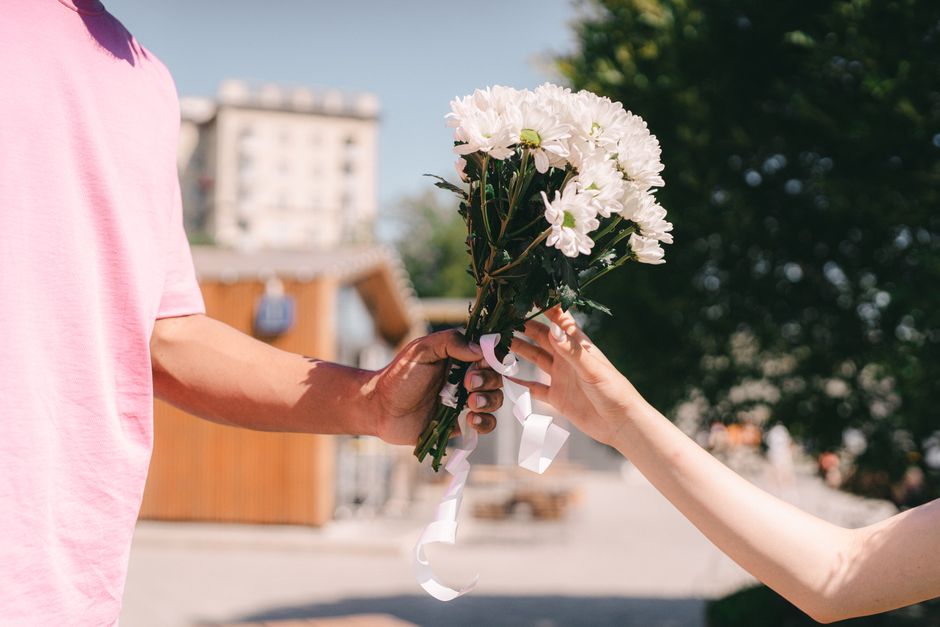 A person wearing a light pink short-sleeved shirt is handing a bouquet of fresh white daisies to another individual outdoors. The bouquet is wrapped in clear plastic and tied with a white ribbon, high