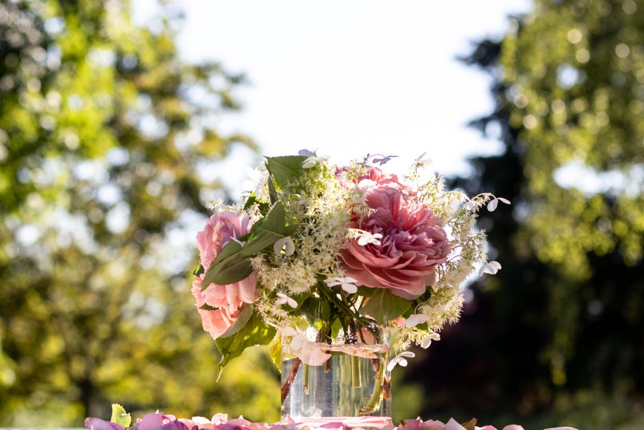 A floral arrangement featuring a bouquet of soft pink roses and white astilbe, accented with green leaves, placed in a transparent glass vase filled with water. The bouquet is set outdoors with natura