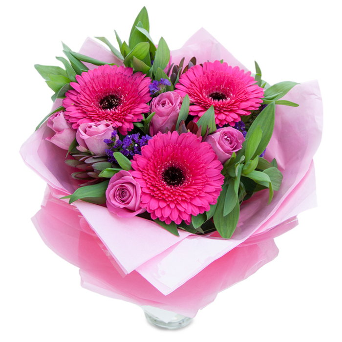 This arrangement reads like a confident note of celebration: three fuchsia-pink gerberas open wide, their layered petals catching light and focusing the eye on their deep centres, while lavender-tinged roses offer a counterpoint of tenderness with petals that roll inward in delicate, velvety curls. Wisps of purple statice punctuate the spaces between larger blooms, bringing texture and a slightly wild, meadow-like charm, and a generous sweep of green foliage - the broad, glossy leaves reminiscent of salal with occasional sprigs of eucalyptus - lends structure and a cool, fresh scent. The composition sits against a stark white background, illuminated by soft, diffuse light that emphasizes the petals' velvety surfaces and the translucent sheen of the wrapping. An inner cushion of pale pink tissue paper frames the stems, overlaid by a sheer fuchsia-pink cellophane that is lightly crinkled and folded to create elegant shadows and highlights; the top rim of a clear glass vase peeks through at the base, suggesting immediacy and readiness for a table or mantlepiece. The visual language here is uplifting and intimate, perfect for a bedside surprise, a birthday table arrangement, or a tender anniversary gift. For residents of the Isle of Dogs it evokes familiar comforts - a bouquet to brighten a flat near South Quay or to bring cheer after a walk through Millwall Park - with a subtle perfume of rose and green notes from gerbera that together feel both fresh and lovingly composed.