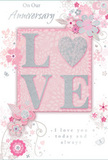 This richly detailed anniversary card is shown as if resting on a tabletop by a window, the soft light revealing every shimmer and line in its design. At its heart, the word LOVE is showcased in prominent, raised lettering, each letter outlined with fine, glittering accents in silver and soft white. The textured surface suggests a satisfying tactile feel, similar to the luxurious ribbon wrapped around a special bouquet delivered in Isle of Dogs. The letters sit on a rectangular blush-pink panel, framed by a slightly paler pink background that spreads to the card's edges. Around this central block, a graceful halo of stylised flowers and curling vines billows outward. The blossoms are drawn with delicate precision in shades of blush, pearl, and pale grey, with a few petals highlighted in shimmering foil-like silver, recalling the elegant, romantic flowers often chosen for local anniversary arrangements. The swirling leaves and tendrils weave in and out of the blooms, creating a light, airy pattern that feels almost like petals drifting on a breeze across the riverside paths near Island Gardens. Between the florals, tiny flourishes and curls add a sense of gentle movement and celebration. The text below or beside the main motif, written in refined script, offers loving anniversary wishes in a tone that feels sincere and enduring. Overall, the card gives the impression of a carefully composed floral sketch brought to life in paper form, the perfect companion to an anniversary bouquet arriving from a nearby Isle of Dogs florist to mark another year of shared love.